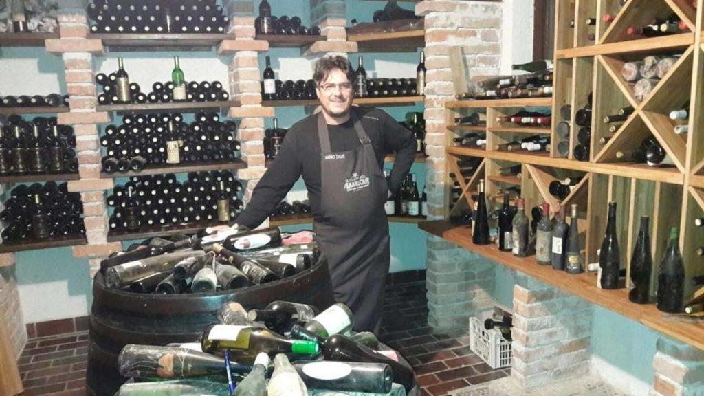 Gostitelj v predpasniku ob sodu v obokani vinski kleti Kluba gurmanov Skaručna, obdan z regali steklenic starega vina